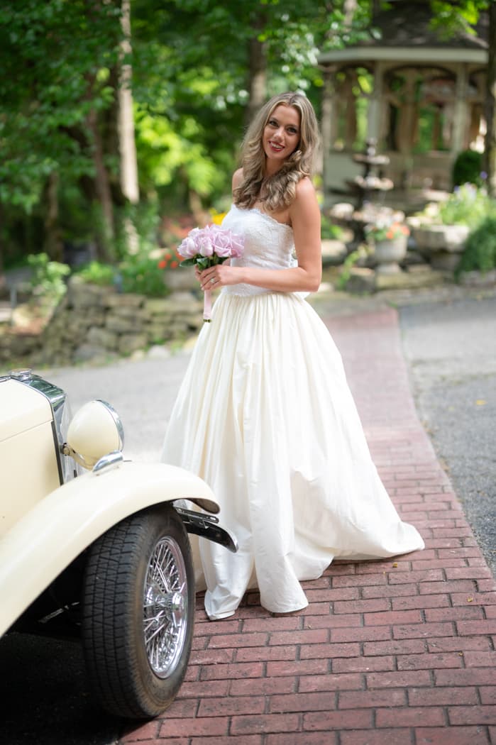 A bride holding pink peonies beside a vintage car on the brick path